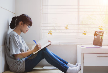 Obraz premium Student learning online study concept: Beautiful Asian girl learning and writing in notebook or homework, sitting short notes, use computer laptop at home for learn of e-learning near window