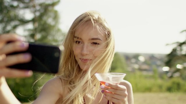 beautiful blonde brown-eyed long-hair girl making selfi with her phone in brown swimsuit and bikini with glass of cocktail a background of nature