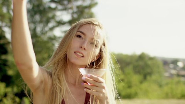 beautiful blonde brown-eyed long-hair girl making selfi with her phone frome up to view in brown swimsuit and bikini with glass of cocktail a background of nature