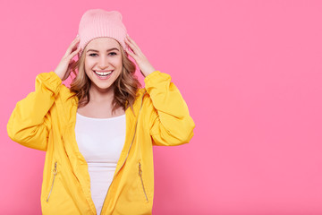 Beautiful hipster teenage girl in bright yellow jacket and pink beanie hat super excited. Attractive cool young woman fashion portrait over pastel pink background.