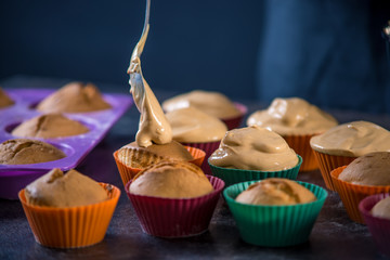 Chef pastry is pouring cream on freshly prepared cupcakes on dark background. Concept of confectionery cooking