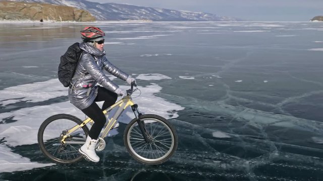 Woman is riding bicycle on the ice. The girl is dressed in a silvery down jacket, cycling backpack and helmet. Ice of the frozen Lake Baikal. The tires on the bicycle are covered with special spikes