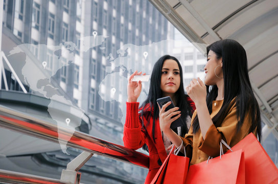 two beautiful asian and caucasian young woman with mobile smart phone, credit card and holding shopping bags together and icon world map network connection diagram, payment, shopping online concept