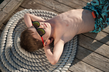 beautiful kid in sunglasses lying on wooden pier with rope under hot summer sun - Powered by Adobe