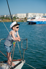 handsome little boy in sunglasses and captain cap on yacht board in sea port in summer vacation day