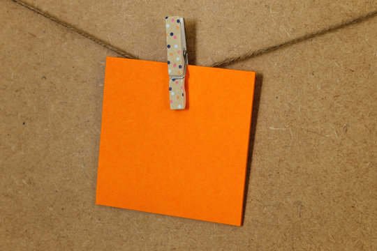 Hoja de papel de color naranja  (posit), sujeto con una pequeña pinza de colore rosa con puntitos blancos, en una cuerda a la pared de corcho. 
