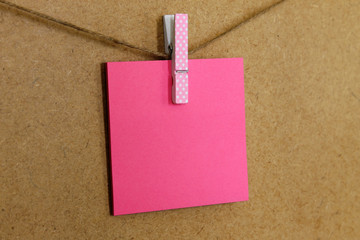 Hoja de papel de color rosa (posit), sujeto con una pequeña pinza de colore rosa con puntitos blancos, en una cuerda a la pared de corcho. 