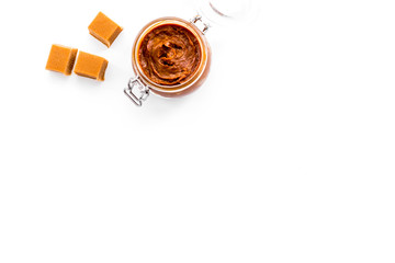 Caramel sauce in glass jar near caramel cubes on white background top view copy space