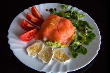 White plate with healthy meal on the black background, in the middle of plate is smoked salmon wrapped around avocado; topped with fish caviar, goat cheese; fresh tomato and lettuce on sides