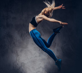 Fototapeta na wymiar Young blonde ballerina in sportswear dances and jumps in a studio. Isolated on a dark background.