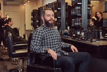 Fototapeta na wymiar Hipster bearded male in a flannel shirt and jeans sitting on a barber chair after shearing in a hairdressing salon.