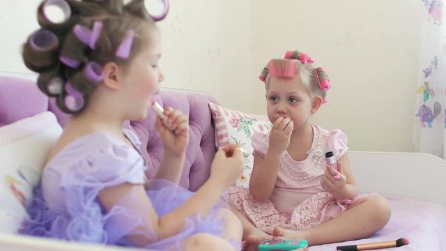 Two little sisters in hair curlers paint their lips and look at each other. 4k