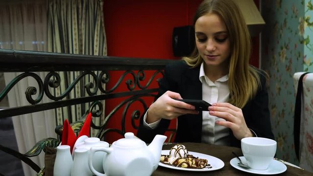 A young girl in a jacket and blouse in a cafe takes a picture of the dessert and selfie