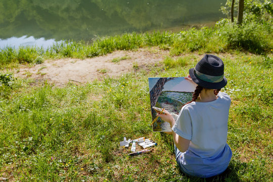 girl-artist draws A river landscape, sitting on the grass, holding a palette and a brush, a canvas, a photo from the back
