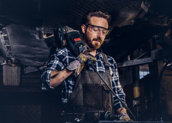 Fototapeta na wymiar Mechanic in a uniform and safety glasses holds an angle grinder while standing under lifting car in a repair garage. 