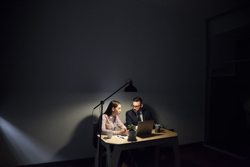 Concentrated colleagues working late hours in dark office room, discussing information, reading news at laptop, analyzing company rates and reports, trying to meet deadline. Hardworking, motivation