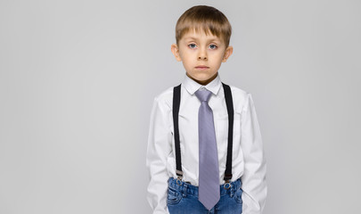 A charming boy in a white shirt, suspenders, a tie and light jeans stands on a gray background