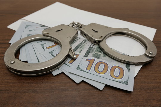 The handcuffs lie on an envelope in which are many hundred dollar bills. Brown wooden background. Concept violation of the law, corruption, financial fraud.
