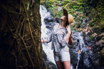 Beautiful young woman standing at the lake near waterfall in white dress and straw hat