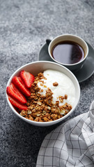 Close up shot of perfect breakfast: crunchy granola with yoghurt and strawberries with a cup of black coffee on gray bakcground.