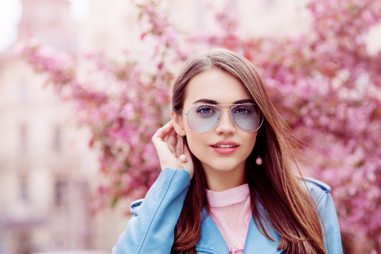 Outdoor close up portrait of young beautiful smiling girl wearing stylish blue color aviator sunglasses, earrings, jacket, posing in street. Spring, summer fashion concept. Copy, empty space for text