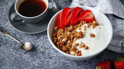 Close up shot of perfect breakfast: crunchy granola with yoghurt and strawberries with a cup of black coffee on gray bakcground.
