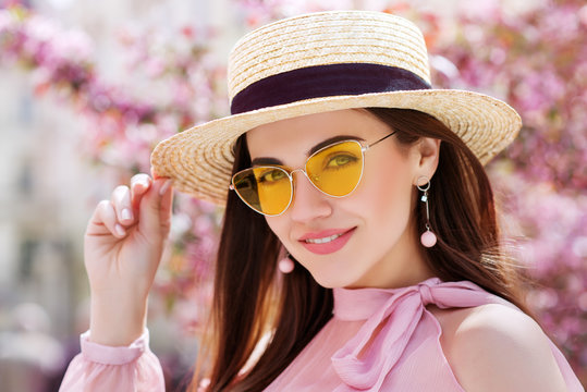 Outdoor close up portrait of young beautiful happy smiling girl wearing trendy yellow color sunglasses, straw boater hat, earrings, off shoulder pink blouse. Spring, summer fashion concept