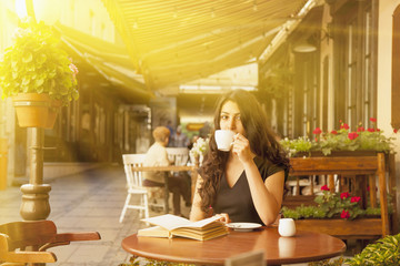 Pretty young woman reading a book and drinking coffee at a sunny day on the terrace of cafe in old European city. (freelancer, relax, travel, education concept)