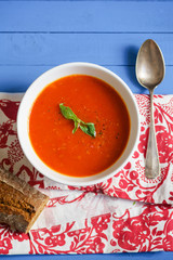 Tomato soup garnished with basil leaves served with rye bread on blue wooden table