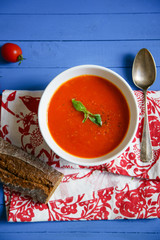 Tomato soup garnished with basil leaves served with rye bread on blue wooden table