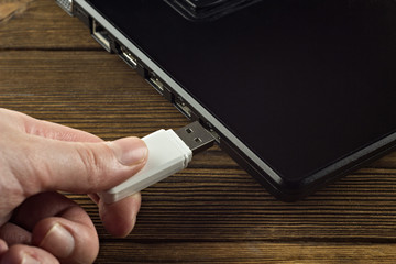 The hand inserts a white USB flash drive into the input of a black laptop, close-up