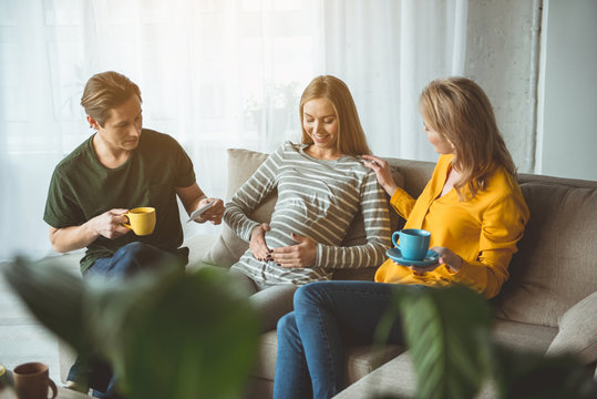 We are waiting for you together. Happy pregnant woman is touching her belly and smiling. Surrogate parents are sitting near and drinking hot beverage 