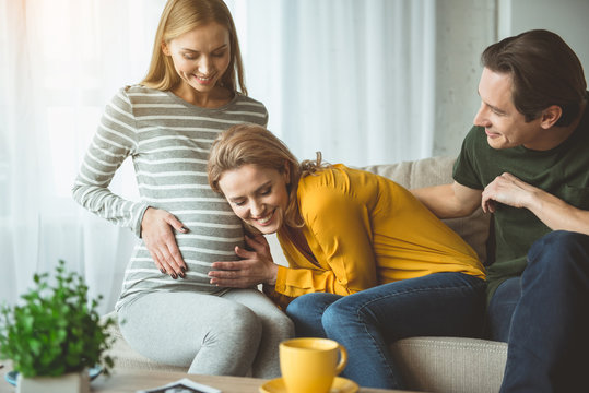 We are waiting for you. Excited young woman is leaning head on female abdomen and hearing to unborn child. Man is sitting near them and smiling. Surrogacy concept 