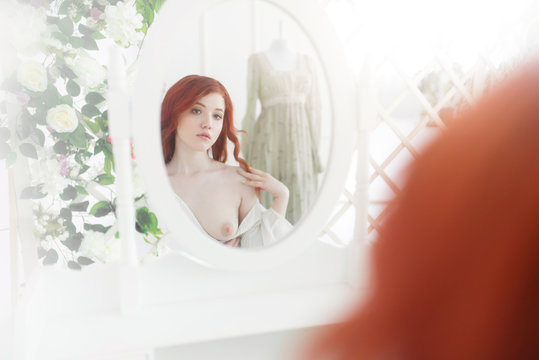 Tender portrait of a young dreamy redhead woman wearing beautiful vintage dress. She is sitting in front of the mirror with naked breast.