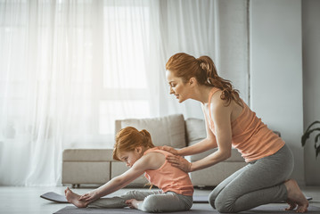 Full length portrait of woman helping her daughter to do stretching exercise on the floor. Lady is smiling and leaning back of her baby girl who looking concentrated