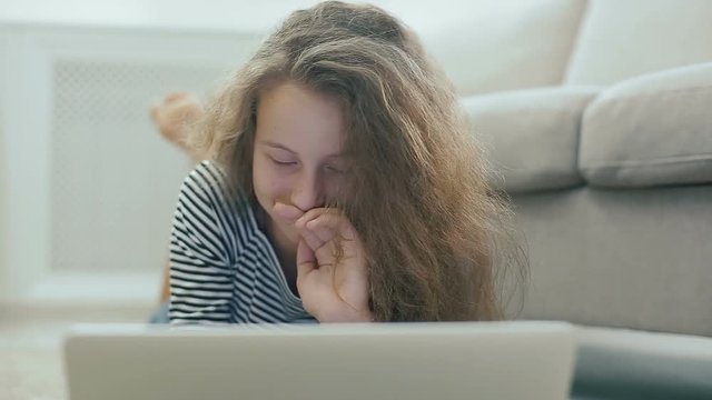 Cute teen girl on a carpet works with laptop. Steadicam shot of happy teen girl near a sofa with laptop. Teen girl uses social networks, reads news, smiles and looks to the camera.