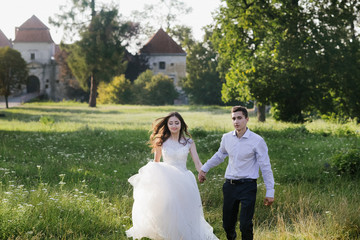 happy wedding couple walking holding hands in evening field on background of old castle. elegant bride and groom embracing. romantic moment. man in suit with bow tie and woman in dress with pearls