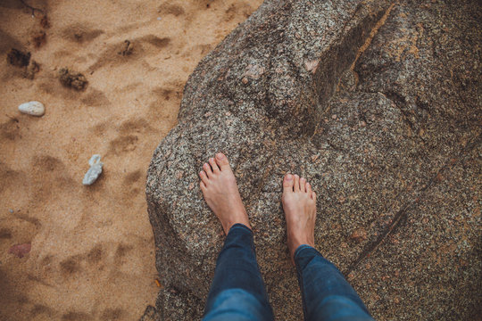 barefooted woman's legs wearing geans standing on the big stone on the beach with yellow sand
