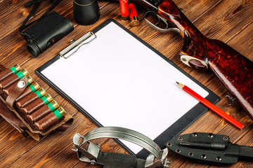 Blank clipboard and pen with hunting equipment on the wooden background.
