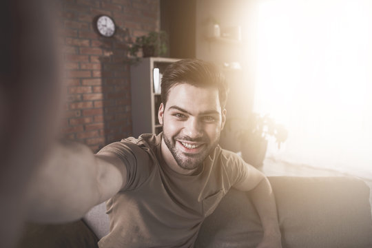 Say cheese. Portrait of young bearded positive man is doing snapshot of himself at home. He is smiling while taking selfie. Sunlight in background