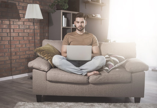 Full-length portrait of young bristled man is sitting in lotus position on comfortable couch. He is holding modern laptop on his knees. Male is looking at camera confidently while laboring at home