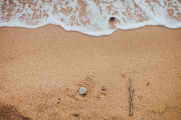 yellow sand and wave with foam from the sea, on the beach lies a stick and a sea stone