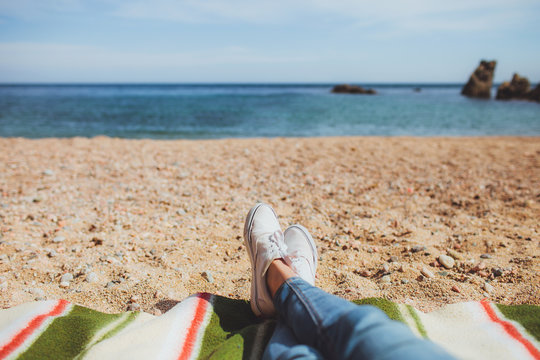 female legs in moccasins and jeans lie on the beach on the veil overlooking the sea, rocks and mountains and a beautiful empty beach in Spain