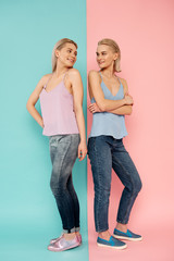 Full length portrait of two sisters standing back to back on blue and pink background. They looking each other with joy