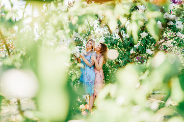 Adorable happy cheerful fabulous twin sisters in different beautiful summer dresses posing outdoor.  Similar cute female models in lilac colorful bushes with bloming flowers in park portrait.  Family.