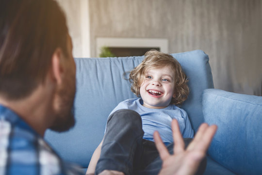 Portrait of laughing child looking at dad while sitting on sofa in room. He gesticulating hands