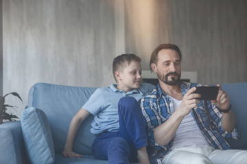 Portrait of cheerful father and satisfied child looking at mobile while situating on sofa. Happy parent and son spending time together concept