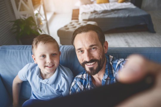 Portrait of beaming man and outgoing kid taking selfie by digital device in room