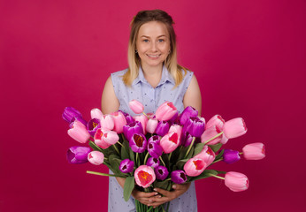 Smiling woman with bunch of flowers in her hands in blue dress. Beautiful woman with bouquet of pink and purple tulips. Summer and spring concept. Isolated on pink background