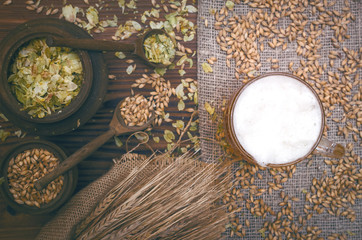 Cold foam beer with malt grain, green hop leaves in the pots and rye ears on burlap cloth on brown wooden table background.
